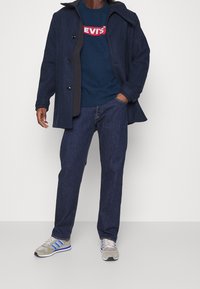 Manteau en laine bleu marine avec de grands boutons noirs, sweat-shirt bleu foncé avec le logo rouge "LEVI'S", jean en denim foncé et baskets grises avec des rayures bleues.
