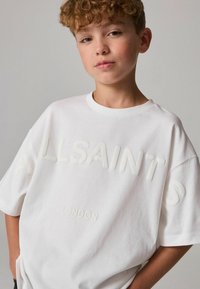 Bílá oversized tričko s embosovaným textem "ALLSAINTS" a "LONDON", kulatý výstřih, krátké rukávy, měkká tkanina, s volným střihem.