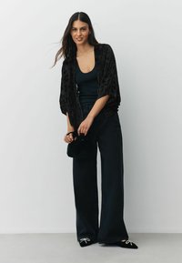 Black velvet kimono with a textured floral design, paired with a black tank top and wide-leg black pants. Completing the look with black shoes featuring rhinestone accents.