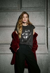 Camiseta gráfica negra con diseño de labios impresos, combinada con un abrigo de piel sintética burdeos y pantalones oscuros de talle alto, frente a una pared de piedra.