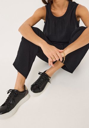 Žena sedí se zkříženýma nohama, má na sobě černý top bez rukávů, černé kalhoty, černé tenisky s bílými podrážkami, zlaté prsteny a náramek.