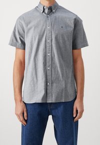 Kortärmad, knappburen skjorta i ljusgrått tyg. Har krage, knappar framtill och en liten logotyp på bröstet. Texturerat material.