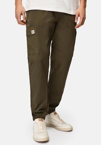 Olivgrüne Cargo-Hosen mit elastischen Bündchen, versehen mit seitlichen Taschen und einem Patch-Logodetail. Getragen mit weißen Sneakers.