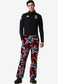 Maglietta nera a mezza zip con logo, abbinata a pantaloni colorati con un motivo astratto rosso, blu e bianco. Fit atletico, con tasche.