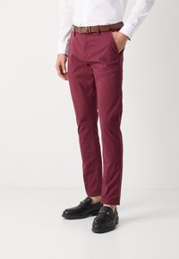 Burgunderfarbene Chinos mit schmaler Passform, aus Baumwolle. Mit frontalen Taschen, einem braunen Gürtel und kombiniert mit schwarzen Slippern.