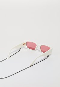 Hvite solbriller med rosa linser, med geometrisk design, gulldetaljer og en svart kjede festet for trygg bruk.