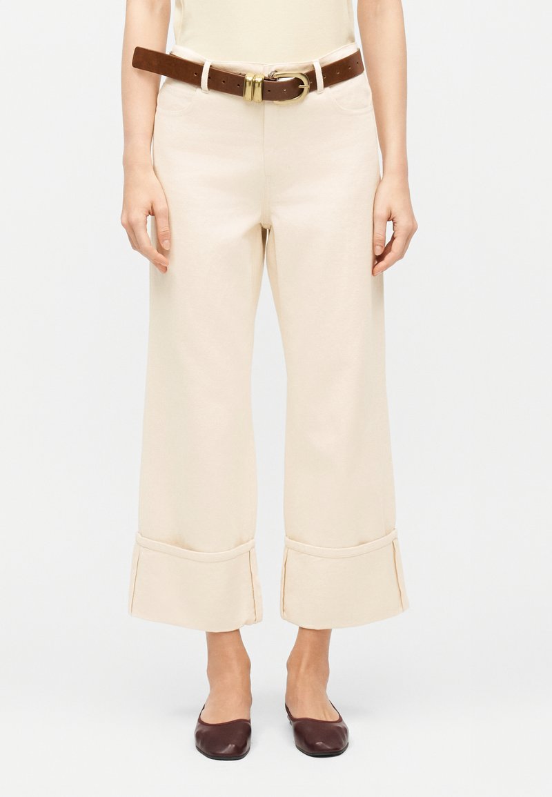 Γυναίκα που φοράει εκρού παντελόνι καμπάνα μέχρι τον αστράγαλο με μεγάλους μανσέτες, καφέ δερμάτινη ζώνη με χρυσή αγκράφα και σκούρα καφέ παπούτσια μπαλέτου.