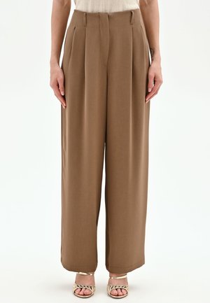 Donna che indossa pantaloni marroni a vita alta e gamba larga e sandali dorati con punta aperta, in piedi di fronte a uno sfondo bianco uniforme.