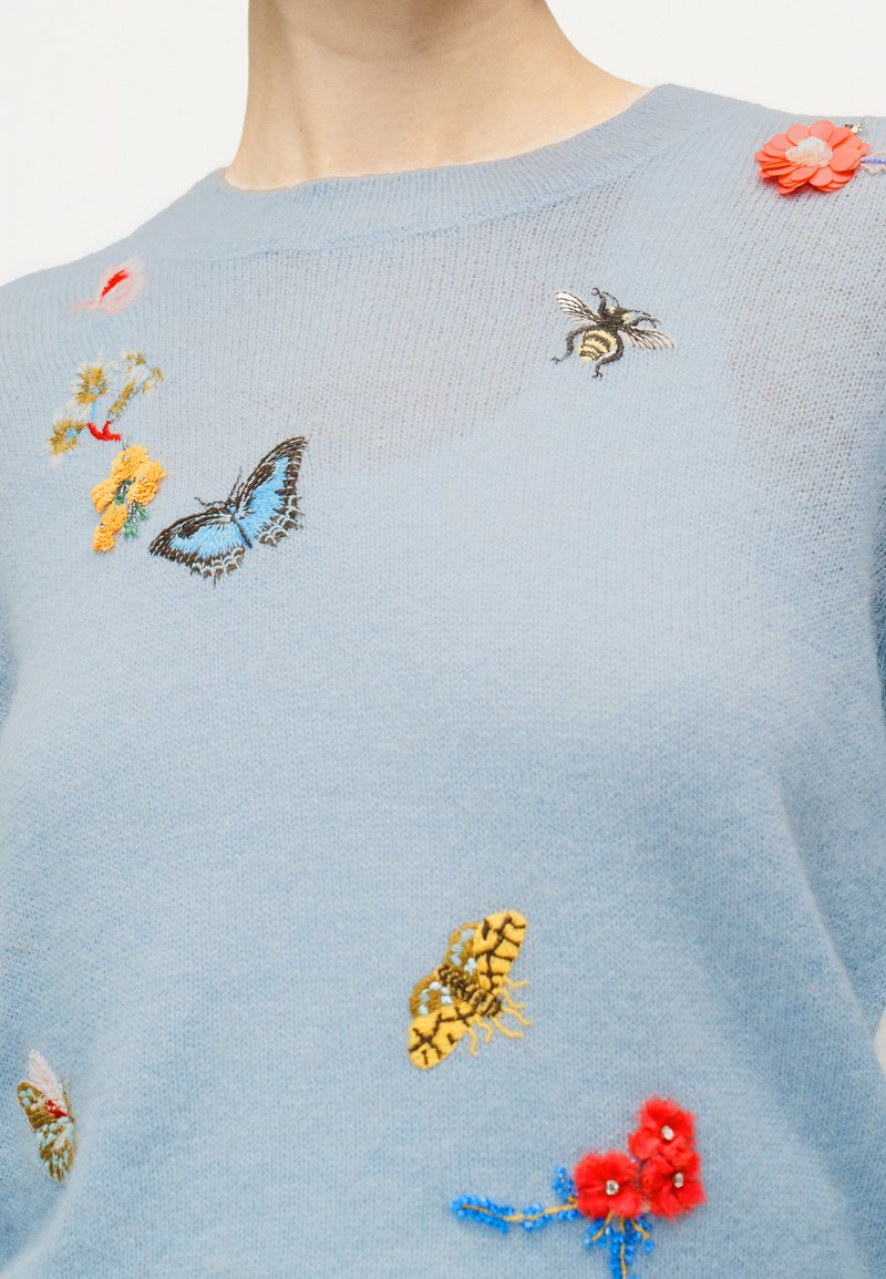 Maglione azzurro chiaro con farfalle, api e fiori ricamati e applicazione di fiore di stoffa rosso vicino alla spalla.