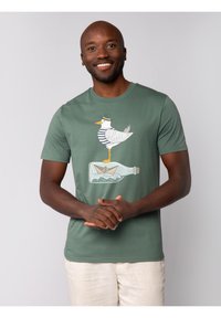 Groene katoenen T-shirt met een grijs en wit cartoon-eend in een gestreept shirt die op een glazen fles staat met een papieren bootje erin.
