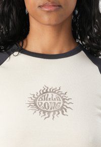 Short-sleeve cream top with dark gray accents, featuring a sun graphic in the center with the text "Silla Pong," ribbed texture.