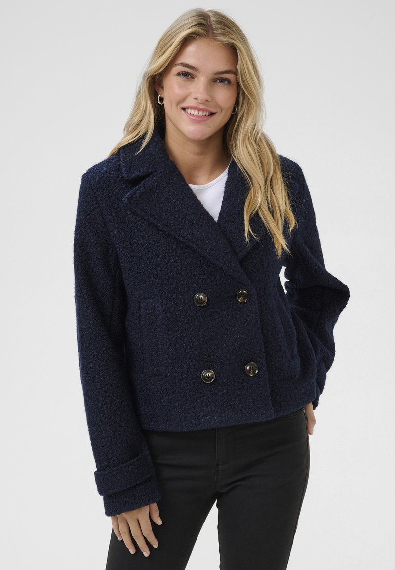 Veste bleu marine croisée en tissu texturé, grand col, deux poches avant et boutons noirs. Stylée avec des manches retroussées.