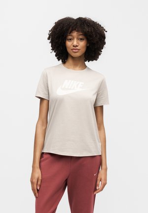 Femme aux cheveux bouclés porte un t-shirt Nike beige clair et un pantalon de survêtement Nike bordeaux, debout devant un fond blanc uni.