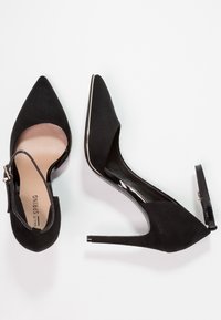 Scarpe con tacco alto in pelle scamosciata nera, con punta affusolata, cinturino alla caviglia e un accento lucido nero lungo il tacco. La suola è liscia e leggermente imbottita.