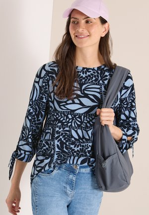 Vrouw met een blauw-zwart top met patroon, lichte spijkerbroek, roze pet en een grijze schoudertas, lacht en kijkt iets naar rechts.