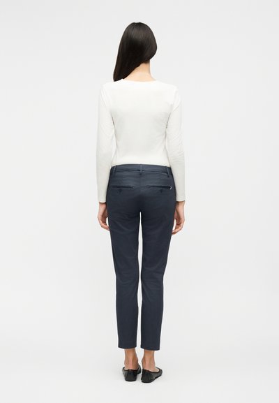 Γυναίκα που στέκεται με την πλάτη, φοράει λευκό μακρυμάνικο τοπ, στενά παντελόνια ναυτικού μπλε χρώματος και μαύρα παπούτσια τύπου slip-on σε απλό φόντο.