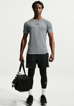 Mand iført grå træningsshirt, sorte shorts over sorte leggings, sorte sneakers, holder en sort sportstaske med et håndklæde mod en ensfarvet baggrund.
