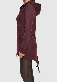 Femme portant un long sweat à capuche zippé bordeaux avec cordons, poches latérales, et des leggings noirs moulants, debout de profil sur fond blanc.