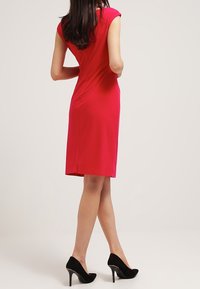 Tajta klänning med kort ärm i en livlig röd färg, mjuk textur, knälång, med en subtil halsringning och söm detajerade i ryggen; parat med svarta klackar.