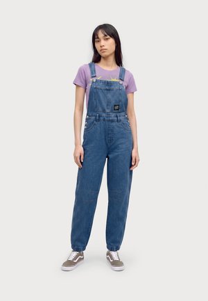 Jeune femme debout portant une salopette en jean bleu, un t-shirt violet avec un motif de soleil et des baskets beiges sur un fond uni clair.