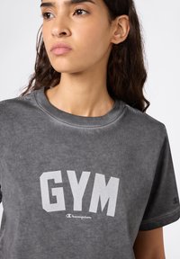Szara bawełniana koszulka z krótkim rękawem, z wyraźnym białym nadrukiem "GYM" oraz małym logo Champion na rękawie. Miękka, gładka tekstura.