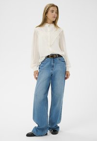 Hvid, semi-gennemsigtig bluse med ruffle-detaljer og knaplukning, parret med løse, lyseblå denim brede bukser og sorte polerede sko.