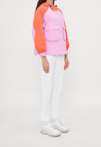 Color-blocked jacket in pink and orange, with a hood, two front pockets, and elastic cuffs. Paired with white pants and light footwear.