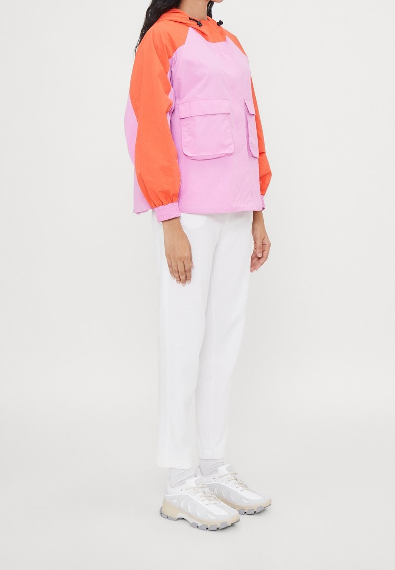 Color-blocked jacket in pink and orange, with a hood, two front pockets, and elastic cuffs. Paired with white pants and light footwear.