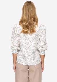 Witte blouse met een klein blauw bloemenpatroon, opgepofte mouwen en geplooide details aan de achterkant, gecombineerd met beige broek met wijde pijpen.
