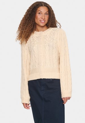 Krämfärgad kabelstickad cardigan med rund halsringning, dekorerad med texturerade mönster och prydliga pom-poms, knäppt med knappar fram.