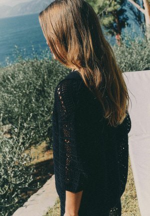 Persoană cu păr lung și castaniu, purtând un pulover tricotat închis la culoare, stând în aer liber lângă vegetație și o întindere de apă.