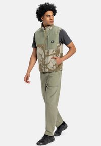 Green and beige fleece vest with zip pockets and a textured upper section, paired with light green pants and black athletic shoes.