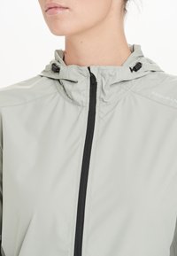 Lichtgrijze windbreaker met capuchon, voorzien van een zwarte rits, verstelbare koord en minimalistisch ontwerp. "ENDURAN" logo op de schouder.