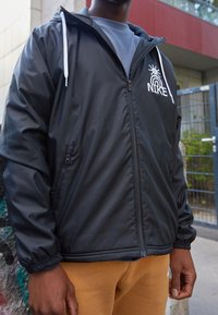 Černá větrovka s zipovým zapínáním, bílými šňůrkami a logem Nike. Disponuje elastickými manžetami a hladkým, lehkým materiálem.