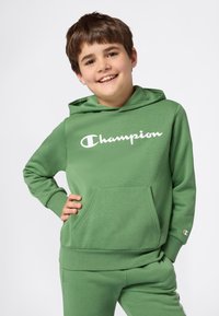 Smilende gutt med kort brunt hår iført grønn Champion hettegenser og matchende bukser, stående med den ene hånden på hoften mot hvit bakgrunn.