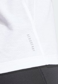 T-shirt de sport blanc en matériau texturé avec un logo "AEROREADY" visible. L'ourlet du t-shirt rencontre des leggings noirs, mettant en valeur le contraste de couleurs.
