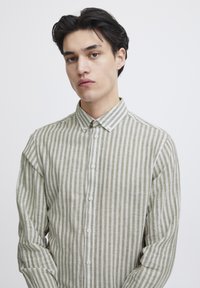 Camisa de manga larga ligera a rayas en verde apagado y blanco, con cuello de botón y cierre frontal con botones. Textura suave, corte relajado.