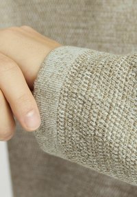 Beige gestrickter Pulloverstoff mit unterschiedlichen Texturen; Nahaufnahme des Ärmelbündchens, das ein glattes, helleres horizontales Streifenmuster zeigt.