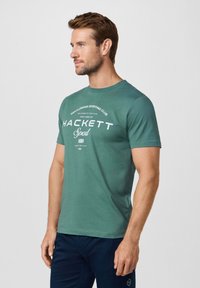 Homme portant un t-shirt teal Hackett Sport et un pantalon bleu marine, debout de profil contre un fond clair uni.