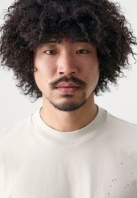 Ljus beige t-shirt med rund halsringning i bomull, med en strukturerad ribbad krage och en subtil prickad mönstring på framsidan.