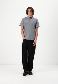 Grå polo t-shirt med stribede krave og logo, parret med løse sorte bukser, båret med sorte sko på en ensfarvet baggrund.