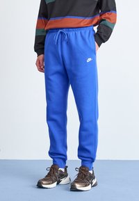 Modré tepláky Nike se stahovacím pasem, žebrovanými lemy na kotnících a bílým logem, doplněné hnědými sportovními botami.