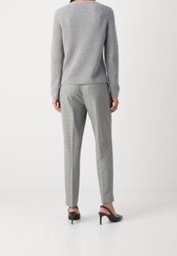 Personne portant un pull gris à côtes, un pantalon carreauté gris et des escarpins à talons à lanières noirs, se tenant sur un sol blanc contre un mur blanc.