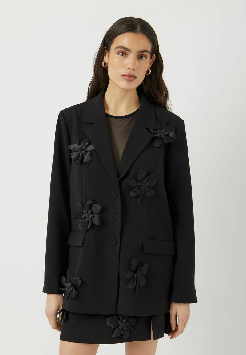 Kvinne iført en svart blazer med store tøyblomsterapplikasjoner over en gjennomsiktig svart topp og matchende svart skjørt med blomsterdetaljer.