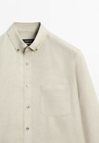 Camicia beige con bottoni, taschino sul petto e colletto con bottoni, mostrata su sfondo bianco.