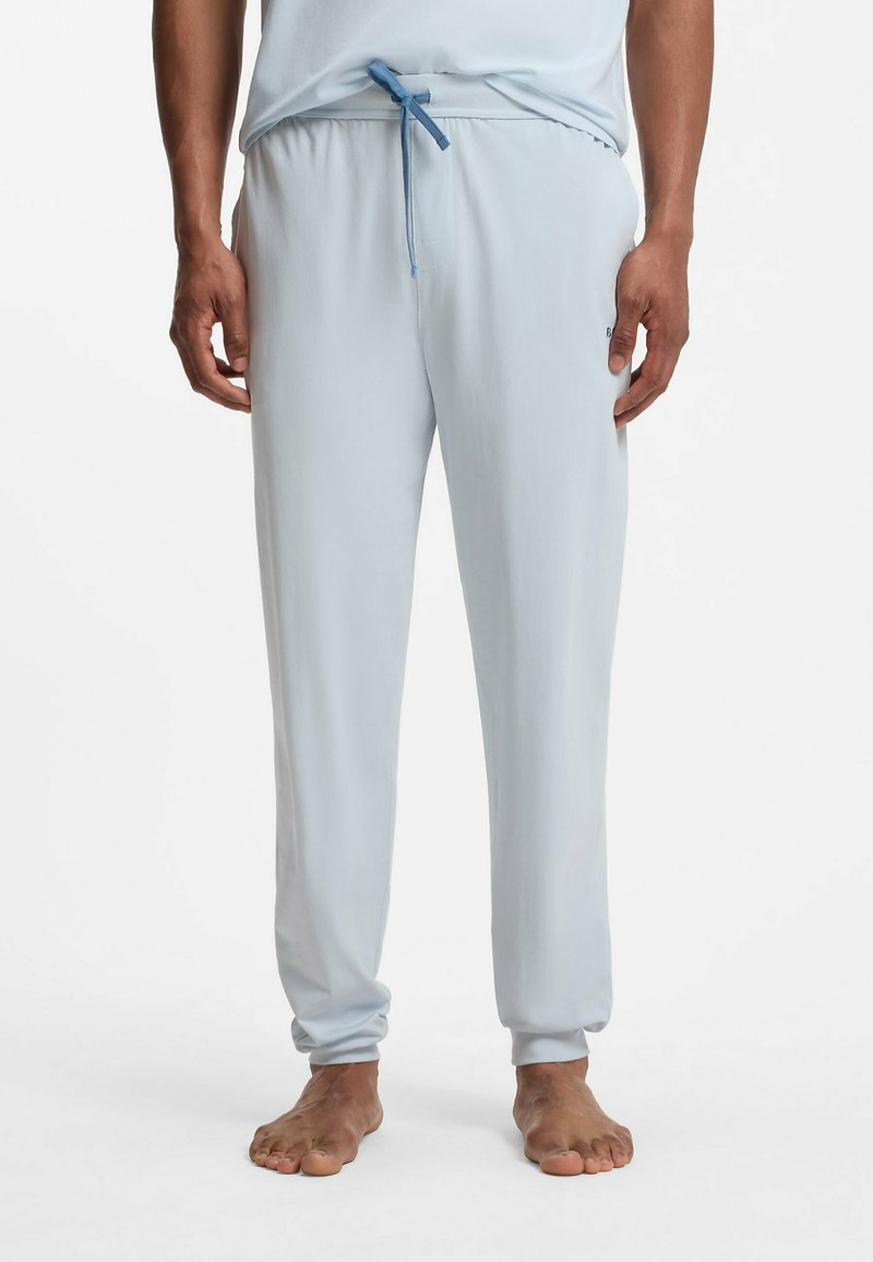 Homme portant un pantalon de jogging bleu clair avec une taille élastique et un cordon, debout pieds nus sur un fond blanc.