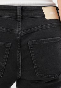 Primo piano di jeans neri in denim sul retro con due tasche e un'etichetta beige cucita sulla cintura, indossati con una maglietta bianca.