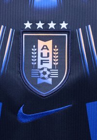 Το έμβλημα της ποδοσφαιρικής ομάδας της Ουρουγουάης με τέσσερα αστέρια πάνω και ένα μπλε λογότυπο της Nike σε μια σκούρα μπλε αθλητική φανέλα με υφή.