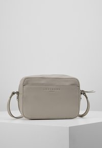 Beżowa torebka typu crossbody z fakturowanej skóry, z zamkiem błyskawicznym, regulowanym paskiem i wytłoczonym logiem marki na przedniej stronie. Kompaktowy prostokątny kształt.