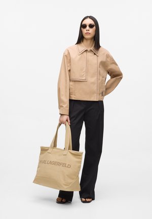Femme portant une veste beige et des lunettes de soleil, pantalon noir, tenant un sac cabas beige Karl Lagerfeld avec des sandales à bout ouvert sur fond blanc.
